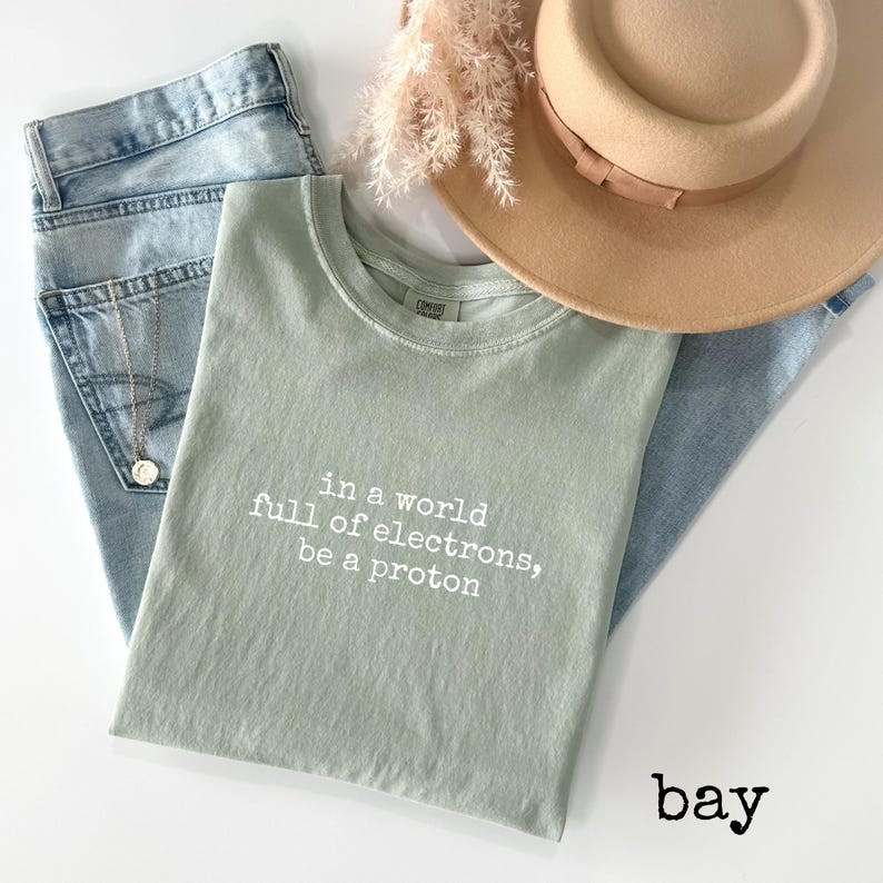 May include: Sage green t-shirt with the text "in a world full of electrons, be a proton" in white. The shirt is laid on top of light blue jeans, with a tan hat and dried flowers in the background.
