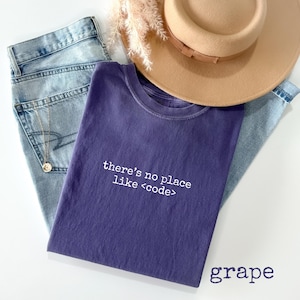May include: A grape-colored t-shirt with the text "there's no place like <code >" in white. The shirt is paired with light blue jeans and a tan hat. A gold necklace is visible.