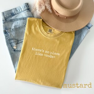 May include: Mustard-colored t-shirt with the text "there's no place like <code >" in white, laid on top of light blue jeans. A tan hat and decorative sprig are also in the image.