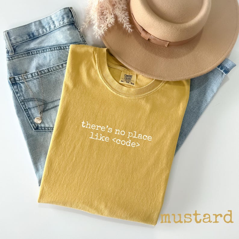 May include: Mustard-colored t-shirt with the text "there's no place like <code >" in white, laid on top of light blue jeans. A tan hat and decorative sprig are also in the image.