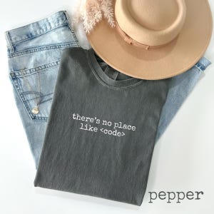 May include: A flat lay featuring a gray t-shirt with the text "there's no place like <code">, light blue jeans, a tan hat, and a necklace. The t-shirt is labeled "pepper".