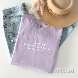 May include: A light purple t-shirt with the text "in a world full of electrons, be a proton" in white. The shirt is paired with light blue jeans, a tan hat, and a gold necklace.
