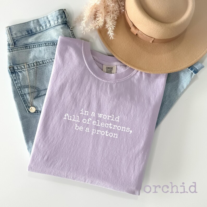 May include: A light purple t-shirt with the text "in a world full of electrons, be a proton" in white. The shirt is paired with light blue jeans, a tan hat, and a gold necklace.