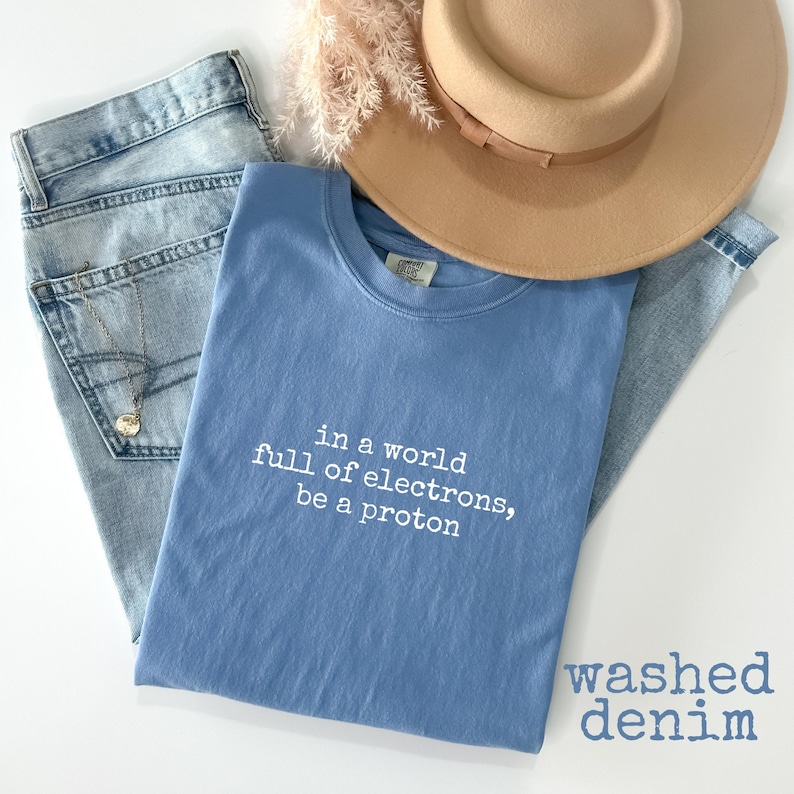 May include: A light blue t-shirt with the text "in a world full of electrons, be a proton" is laid on top of light blue denim jeans. A tan hat and a necklace with a pendant are also in the image. The words "washed denim" are in the bottom right corner.