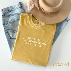 May include: Mustard-colored t-shirt with the text "in a world full of electrons, be a proton." The shirt is paired with light-wash denim jeans and a tan fedora hat. A necklace with a pendant is visible in the jeans pocket.