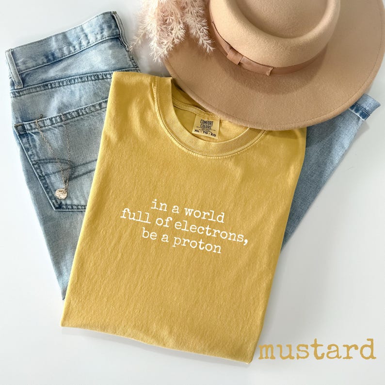 May include: Mustard-colored t-shirt with the text "in a world full of electrons, be a proton." The shirt is paired with light-wash denim jeans and a tan fedora hat. A necklace with a pendant is visible in the jeans pocket.