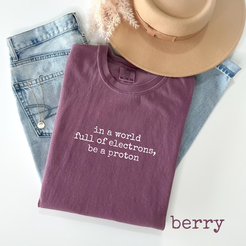 May include: A berry-colored t-shirt with the text "in a world full of electrons, be a proton" in white. The shirt is paired with light blue jeans and a tan hat.