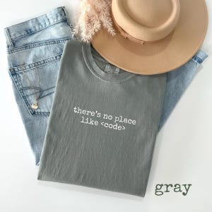 May include: A gray t-shirt with the text "there's no place like <code >" in white, laid on top of light blue jeans. A tan hat and a gold necklace are also visible. The word "gray" is printed in the bottom right corner.