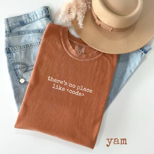 May include: An orange-brown t-shirt with the text "there's no place like <code >" in white. The shirt is laid on top of light blue jeans, a tan hat, and a gold necklace. A sprig of dried flowers is also present.