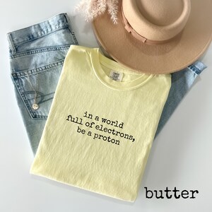 May include: A light yellow t-shirt with the text "in a world full of electrons, be a proton" in black. The shirt is folded and rests on a pair of blue jeans, with a beige hat and a necklace visible.