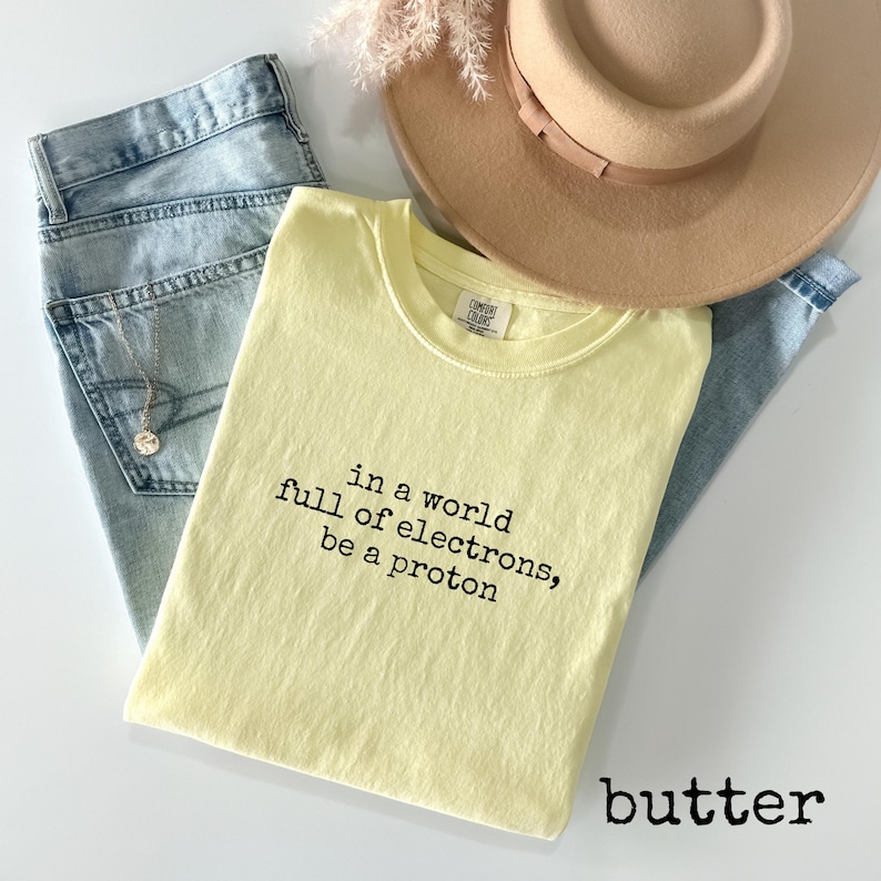 May include: A light yellow t-shirt with the text "in a world full of electrons, be a proton" in black. The shirt is folded and rests on a pair of blue jeans, with a beige hat and a necklace visible.