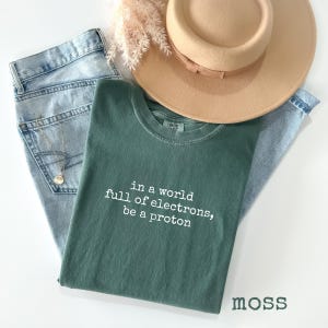 May include: A casual fashion ensemble featuring a moss green t-shirt with the text "in a world full of electrons, be a proton", light wash denim jeans, a tan hat, and a gold necklace. The word "moss" is printed at the bottom.