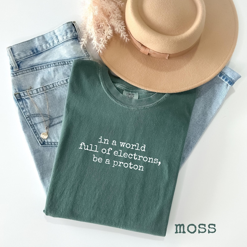 May include: A casual fashion ensemble featuring a moss green t-shirt with the text "in a world full of electrons, be a proton", light wash denim jeans, a tan hat, and a gold necklace. The word "moss" is printed at the bottom.