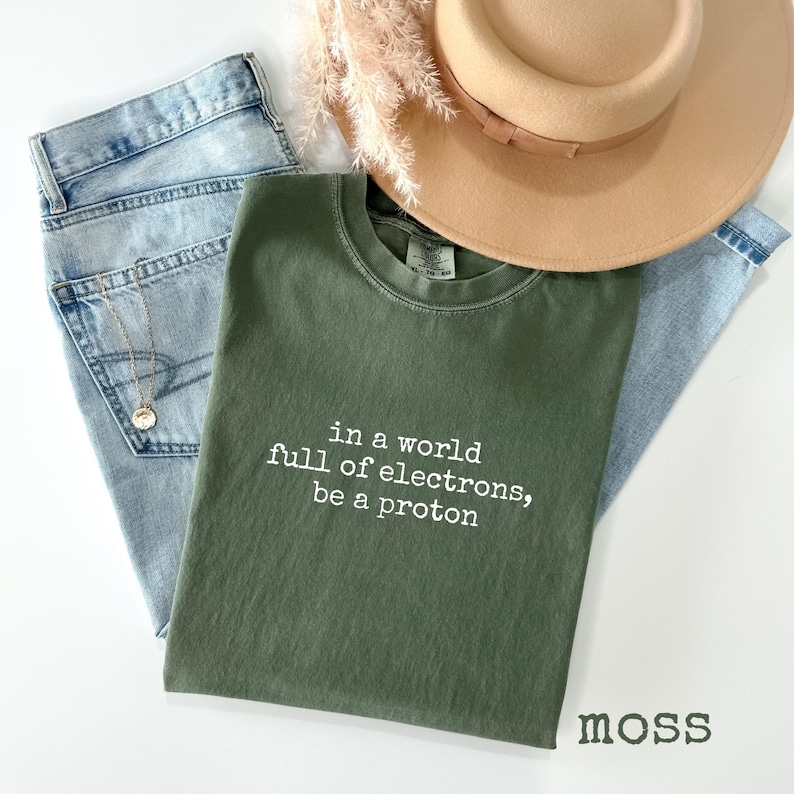 May include: An olive green t-shirt with the text "in a world full of electrons, be a proton" in white. The shirt is paired with light wash denim jeans, a gold necklace, and a tan hat. Dried flowers are also present.