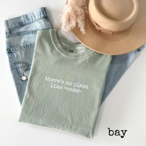 May include: A sage green t-shirt with the text "there's no place like <code >" in white. The shirt is laid on top of light blue jeans, with a gold necklace, a tan hat, and dried flowers. The word "bay" is written in black.