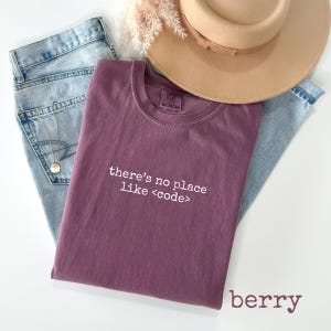 May include: A berry-colored t-shirt with the text "there's no place like <code"> is laid on top of light blue jeans. A tan hat and a gold necklace are also in the image. The word "berry" is written in the bottom right corner.