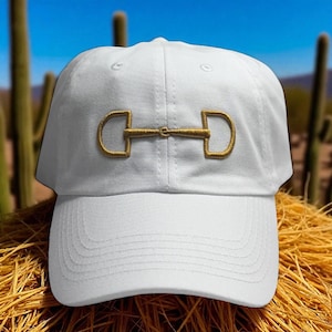 May include: White baseball cap with a gold-colored horse bit embroidered on the front. The cap has a curved brim and a structured crown. The background features a desert landscape with cacti and a blue sky.