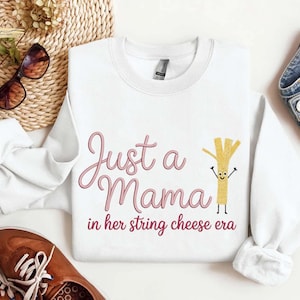 Puede incluir: Sudadera blanca con el texto "Just a Mama in her string cheese era" en escritura rosa. También aparece un personaje de dibujos animados de queso en hebras. La sudadera se combina con zapatos marrones y vaqueros azules.