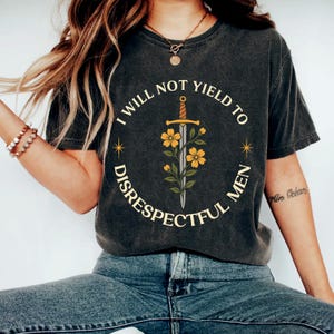 Puede incluir: Camiseta gris oscuro con el texto "I WILL NOT YIELD TO DISRESPECTFUL MEN" en un diseño circular. El diseño incluye una espada, flores amarillas y estrellas. La camiseta es de estilo casual.