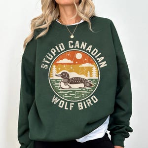 May include: Dark green sweatshirt with a circular graphic featuring a loon on water, trees, and a sunset. The text reads "STUPID CANADIAN WOLF BIRD" in a distressed font. The sweatshirt has a crew neck and long sleeves.