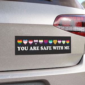Puede incluir: Adhesivo rectangular negro para parachoques con el texto "YOU ARE SAFE WITH ME" en blanco. Encima del texto hay formas de corazón coloridas que representan varias banderas del orgullo LGBTQ+. La pegatina está adherida a la parte trasera de un coche plateado.