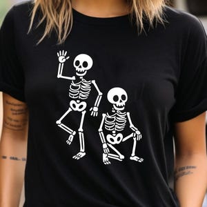 May include: Black t-shirt featuring a white graphic of two skeletons. One skeleton is waving, and the other is kneeling. The design is simple and bold, perfect for Halloween or everyday wear.