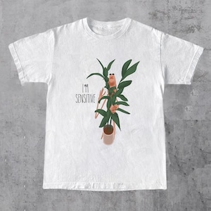 以下が含まれることがあります： 白いTシャツ。植物の体を持つ人が鉢植えの植物を持っている、気まぐれなイラストが特徴です。「I'M SENSITIVE」の文字がその上に印刷されています。デザインは緑、茶色、ベージュの色合いです。