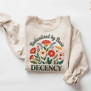 Peut inclure: Sweat-shirt crème avec un motif floral et le texte "Radicalized by Basic Decency". Le motif présente des fleurs rouges, oranges et jaunes avec des feuilles vertes.