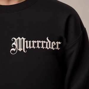 Peut inclure: Sweat-shirt noir avec le mot "Murrrder" brodé en lettres gothiques crème sur la poitrine. Le texte brodé ressort sur le tissu sombre. Le sweat-shirt a un col rond et des manches longues.
