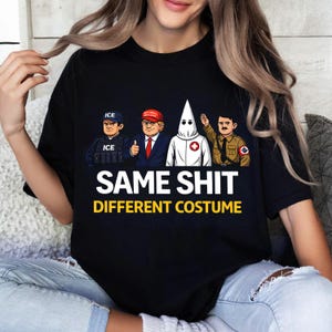 Könnte beinhalten: Schwarzes T-Shirt mit einer Grafik, die vier Cartoon-Figuren zeigt: einen ICE-Agenten, einen Mann mit roter Mütze, einen Ku-Klux-Klan-Mann und einen Mann in Nazi-Uniform. Der Text "SAME SHIT DIFFERENT COSTUME" ist darunter gedruckt.