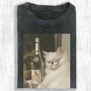May include: Dark gray t-shirt featuring a graphic of a white cat wearing round glasses, next to a liquor bottle. The bottle has a silver cap and a label with text. The cat is in a vintage-style image.