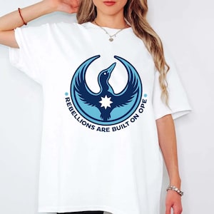 Peut inclure: T-shirt blanc avec un motif bleu et blanc d'un oiseau dans un cercle. Le texte "REBELLIONS ARE BUILT ON OPE" est imprimé autour de l'oiseau. Le t-shirt a des manches courtes et une coupe ample.