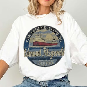 May include: White t-shirt featuring a circular graphic with a red and white ship illustration. The graphic includes the text "THE LEGEND LIVES ON" and "Edmund Fitzgerald" with "NOV. 10th 1975 - LAKE SUPERIOR" below. The shirt is a casual, oversized style.