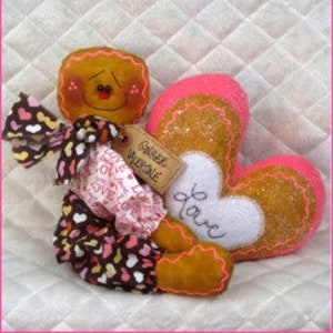 May include: A gingerbread person doll with a pink and white heart-shaped pillow. The pillow has the word "love" embroidered on it. The doll is wearing a pink and brown dress with white hearts. The doll has a tag that says "Gingerbread Valentine".