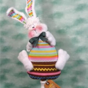 Może przedstawiać: Biały pluszowy królik z różowymi policzkami i zielonym krawatem. Królik nosi prążkowaną koszulę i ma brązowo-białą prążkowaną spódnicę. Królik siedzi na białym cokole z etykietą, na której napisano "The Bunny Hutch".