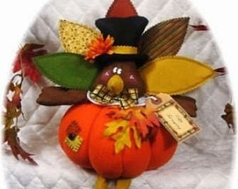 Puede incluir: Un pavo de Acción de Gracias de fieltro con cuerpo de calabaza, sombrero marrón y negro y patas amarillas. El pavo lleva una bufanda de cuadros y una etiqueta que dice "Feliz Día de Acción de Gracias".