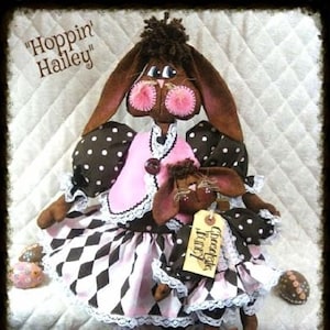 May include: Two brown bunny dolls with pink accents. The larger doll is wearing a pink vest, a black and white checkered skirt, and white lace trim. The smaller doll is wearing a pink dress with black and white checkered trim and white lace. The larger doll has a tag that says "Chocolate Bunny".