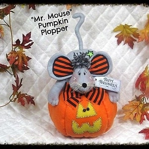 May include: A gray mouse with black and orange striped ears and a tail is sitting inside a carved orange pumpkin. The pumpkin has a happy Halloween tag attached to it. The mouse is wearing a black and orange striped scarf. The background is a white quilted fabric.