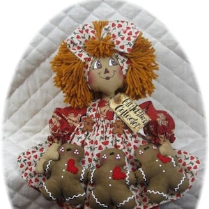 Könnte beinhalten: Eine handgemachte Stoffpuppe mit einem roten und weißen Herzmusterkleid, braunen Garnhaaren und einer roten Schleife. Die Puppe hält drei Lebkuchenmänner mit roten Herzen und weißem Zuckerguss. Die Puppe hat ein Schild mit der Aufschrift "Lebkuchen-Sammler".