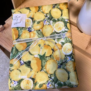 May include: A folded fabric with a lemon pattern, tied with a white ribbon. The fabric features yellow lemons, green leaves, and small purple flowers. A tag indicates dimensions of 55 inches by 40 inches.