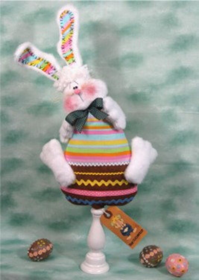 May include: A white stuffed bunny with pink cheeks and a green bow tie. The bunny is wearing a striped shirt and has a brown and white striped skirt. The bunny is sitting on a white pedestal with a tag that says "The Bunny Hutch".