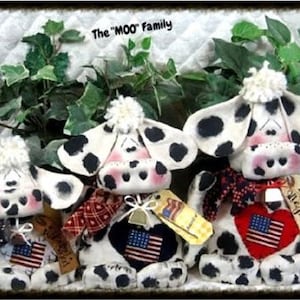 Op de afbeelding: Handgemaakte koeienknuffels met zwarte vlekken, witte lichamen en roze accenten. Elke koe heeft een patroon sjaal en een label met een Amerikaanse vlag. De tekst "The 'MOO' Family" staat bovenaan.