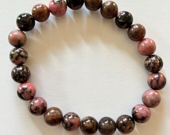 May include: A bracelet made of polished, round beads in pink, brown, and black. The beads are strung together to form a circular shape. The stones have a marbled effect, adding visual interest to the jewellery. The bracelet is suitable for a wrist.