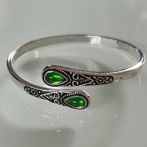 May include: Silver bangle bracelet with two green gemstone accents. The bracelet is open and has a decorative design.