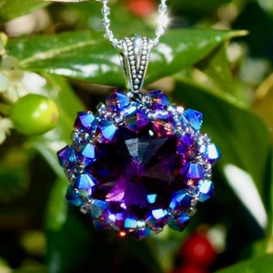 May include: A pendant necklace featuring a large, faceted purple gemstone surrounded by smaller, iridescent blue and purple crystals. The pendant hangs from a delicate silver chain, set against a backdrop of green foliage.