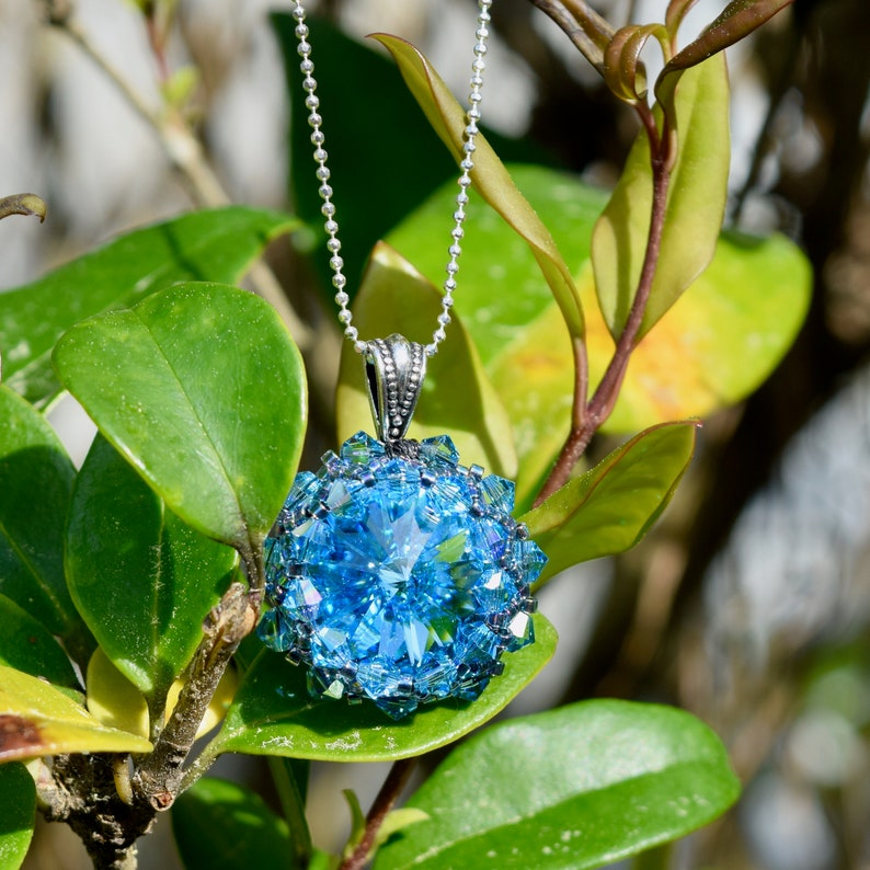 Op de afbeelding: Een close-up van een hangerketting met een grote, facetgeslepen, lichtblauwe kristal. De kristal is omgeven door kleinere, bijpassende blauwe kristallen. De hanger hangt aan een fijne zilveren ketting, tegen een achtergrond van groene bladeren.