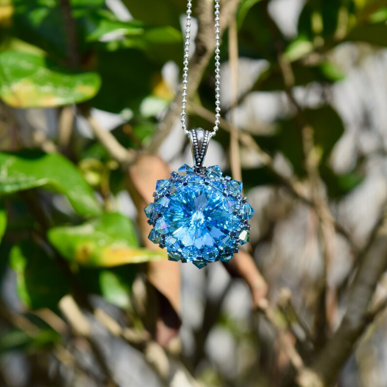 Op de afbeelding: Een opvallende blauwe kristallen hangerketting. De hanger heeft een grote, facetgeslepen, lichtblauwe kristal, omringd door kleinere, veelzijdige kristallen. Hij hangt aan een fijne zilveren ketting, wat contrasteert met de groene bladeren op de achtergrond.