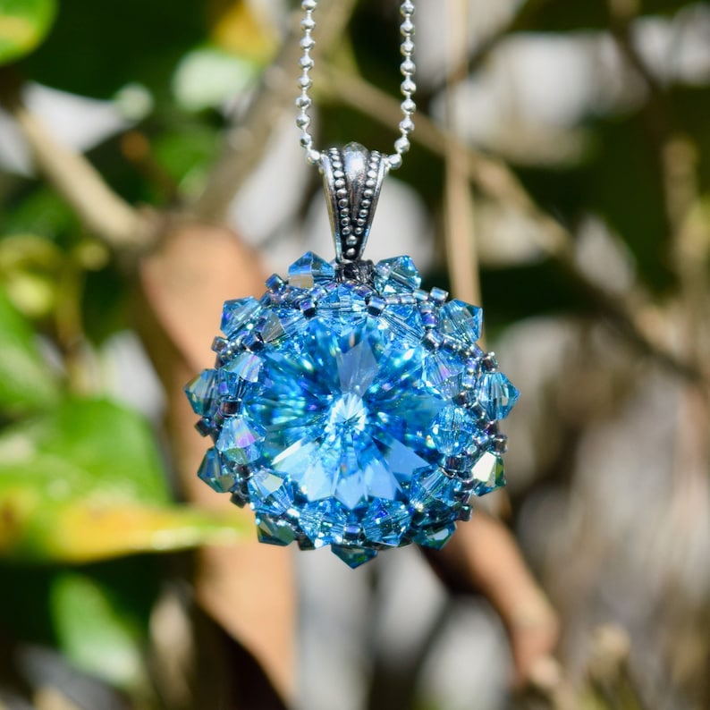 Op de afbeelding: Een opvallende blauwe hangerketting. De hanger heeft een grote, facetgeslepen, lichtblauwe kristal in het midden, omringd door kleinere, glinsterende blauwe kristallen. De hanger hangt aan een fijne zilveren ketting, wat een prachtig sieraad cre&euml;ert.