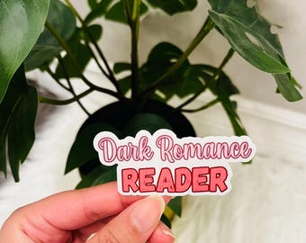 May include: A die-cut sticker with a white border featuring the text Dark Romance in a pink cursive font on the top line and the word READER in a bold, blocky, textured red font on the bottom line, presented against a green plant background.