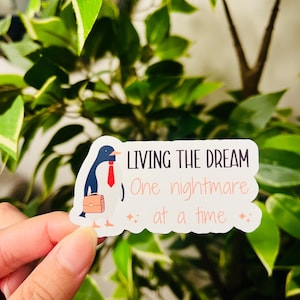 May include: A white sticker featuring a cartoon penguin in a red tie and a briefcase. The sticker reads "LIVING THE DREAM" above "One nightmare at a time" with star accents.